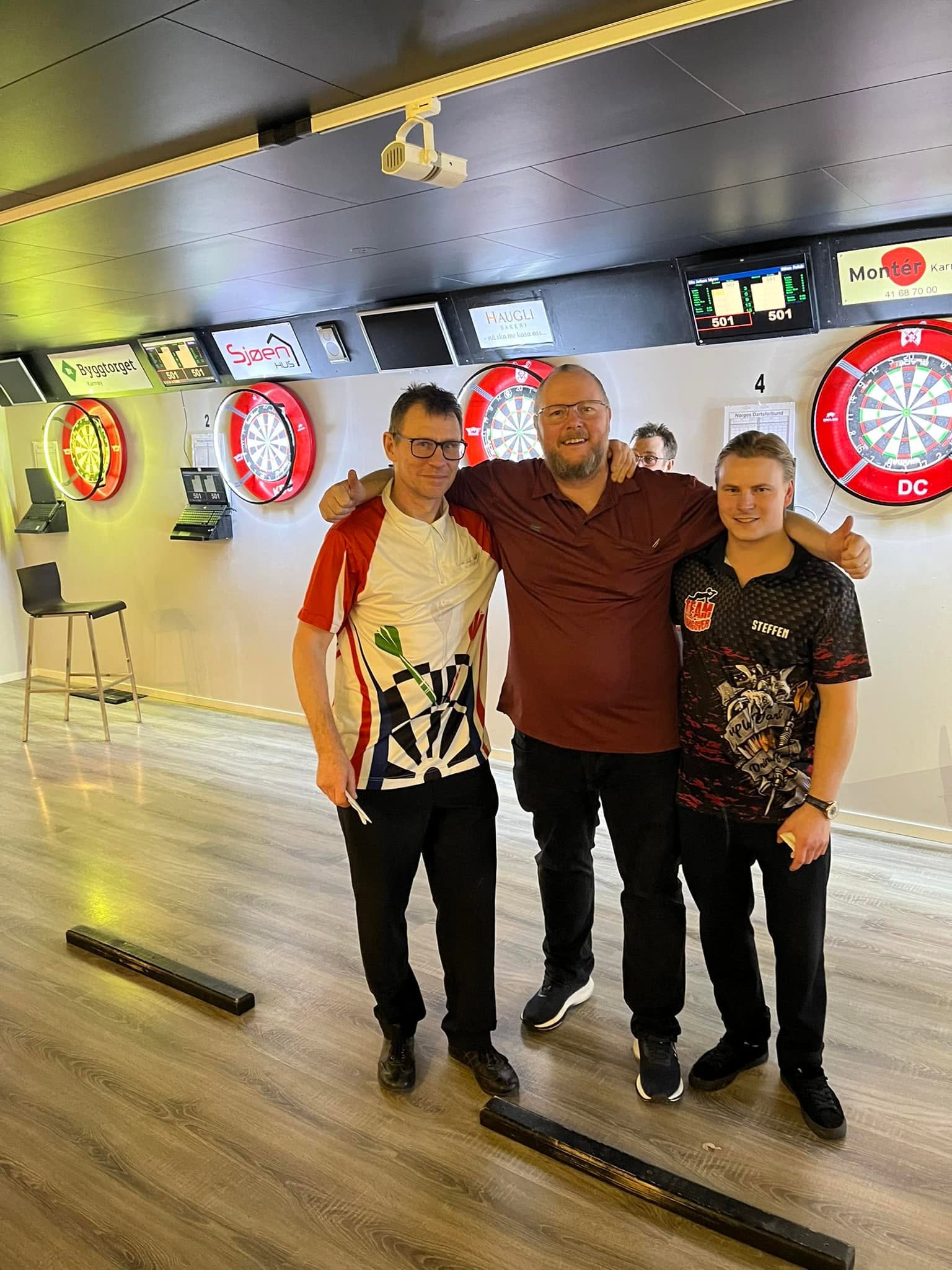 Tre smilende deltakere foran dartbanene